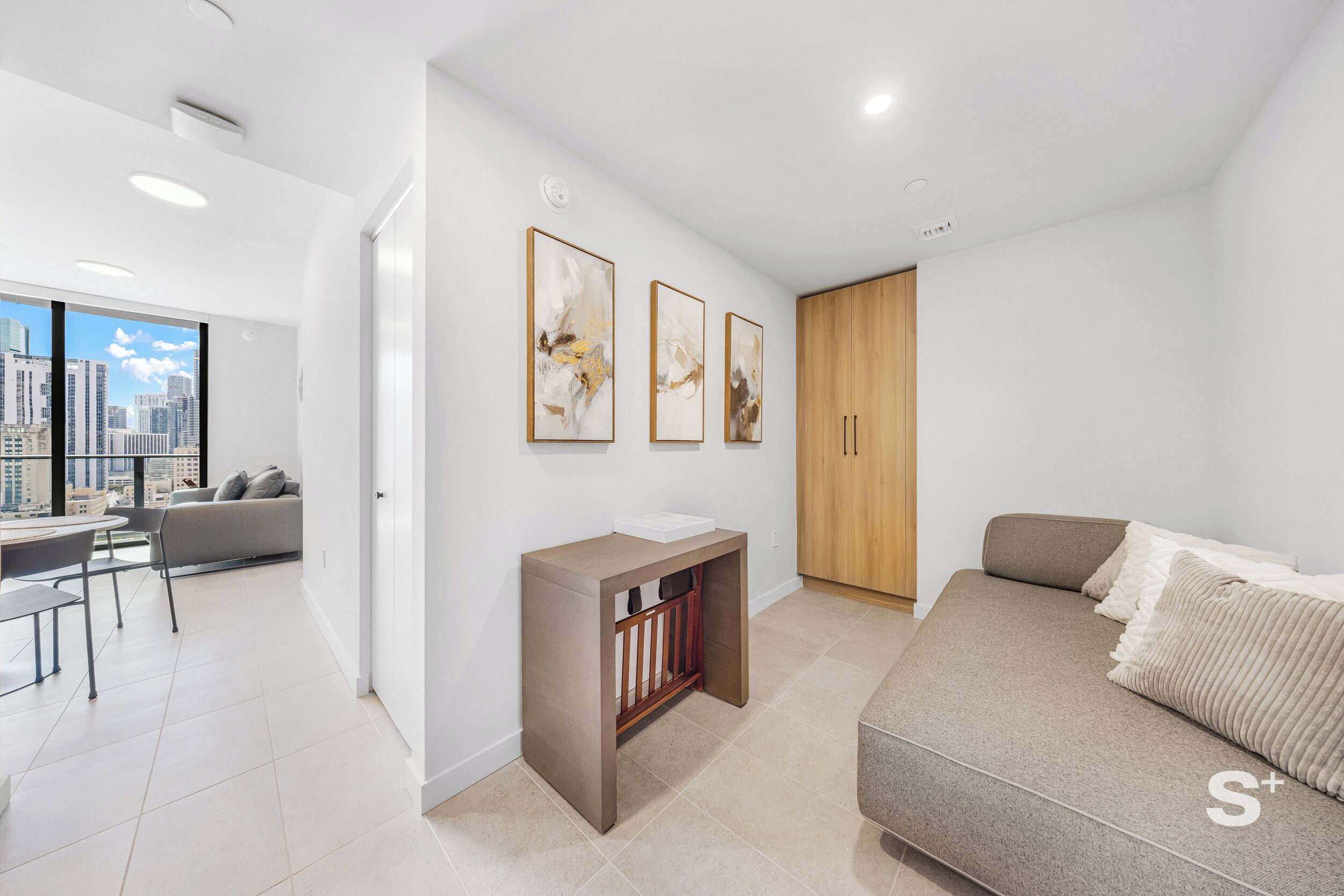 Uma sala de estar moderna com sofá, uma mesinha e um berço. Há três quadros abstratos na parede e um guarda-roupa de madeira.