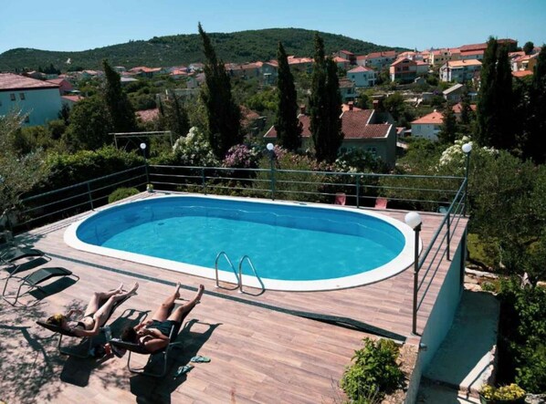 Une piscine dotée d’une terrasse en bois et de chaises longues, entourée de verdure et offrant une vue sur un quartier résidentiel situé sur une colline.