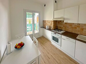 A kitchen with white cabinets, a gas stove, an oven, and a dining table with a bowl of fruit. - Magnificent Apartment in park residence in 5 min from the seaside (Nice)