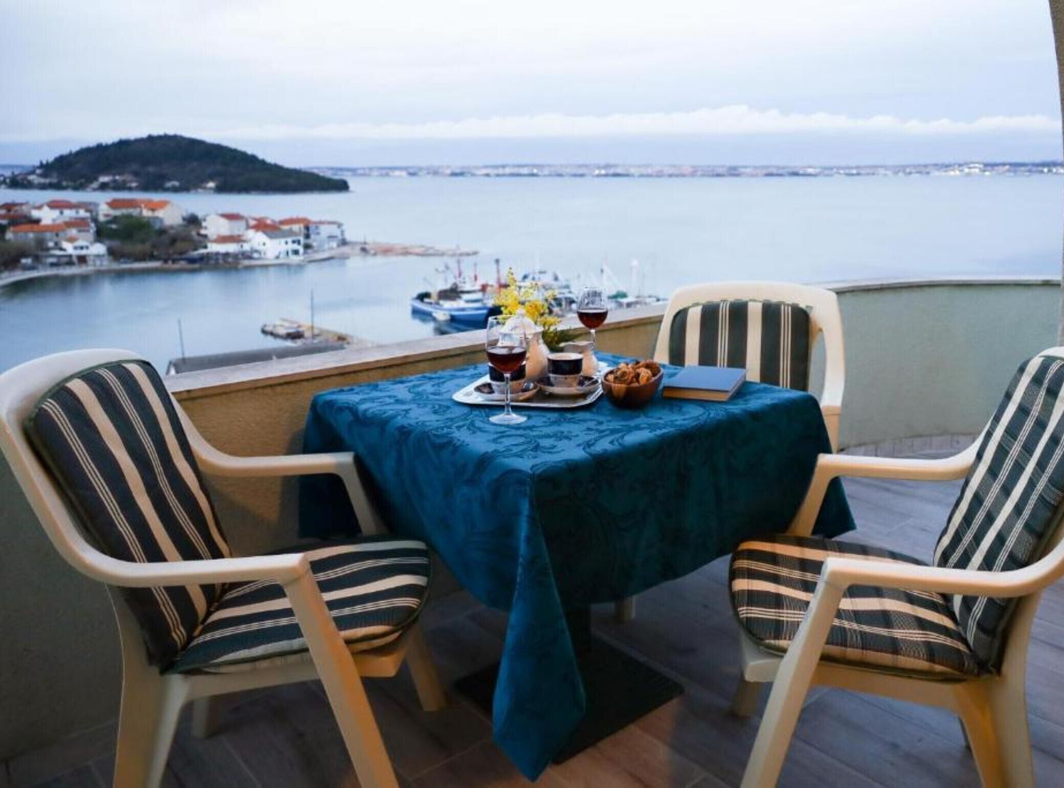 A balcony table with two striped chairs, a tablecloth, and a view of a body of water with boats and a distant island.