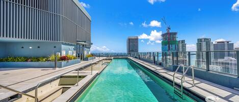 Une piscine sur le toit offrant une vue sur la ville, un bâtiment moderne aux lignes verticales et un ciel d’un bleu limpide.