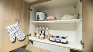 Una cocina con armarios de madera, un paño de cocina colgado y estantes repletos de aceite de cocina, salsa de soja, salsa de pescado, vinagre, sal, pimienta, crema, café y azúcar.