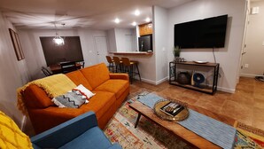 Ein Wohnzimmer mit einer Couch, einem Couchtisch, einem Fernseher und einer Küchenzeile im Hintergrund.