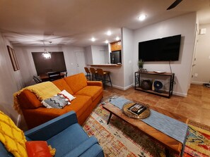 Ein Wohnzimmer mit einer Couch, einem Couchtisch, einem Fernseher und einer Küchenzeile im Hintergrund.