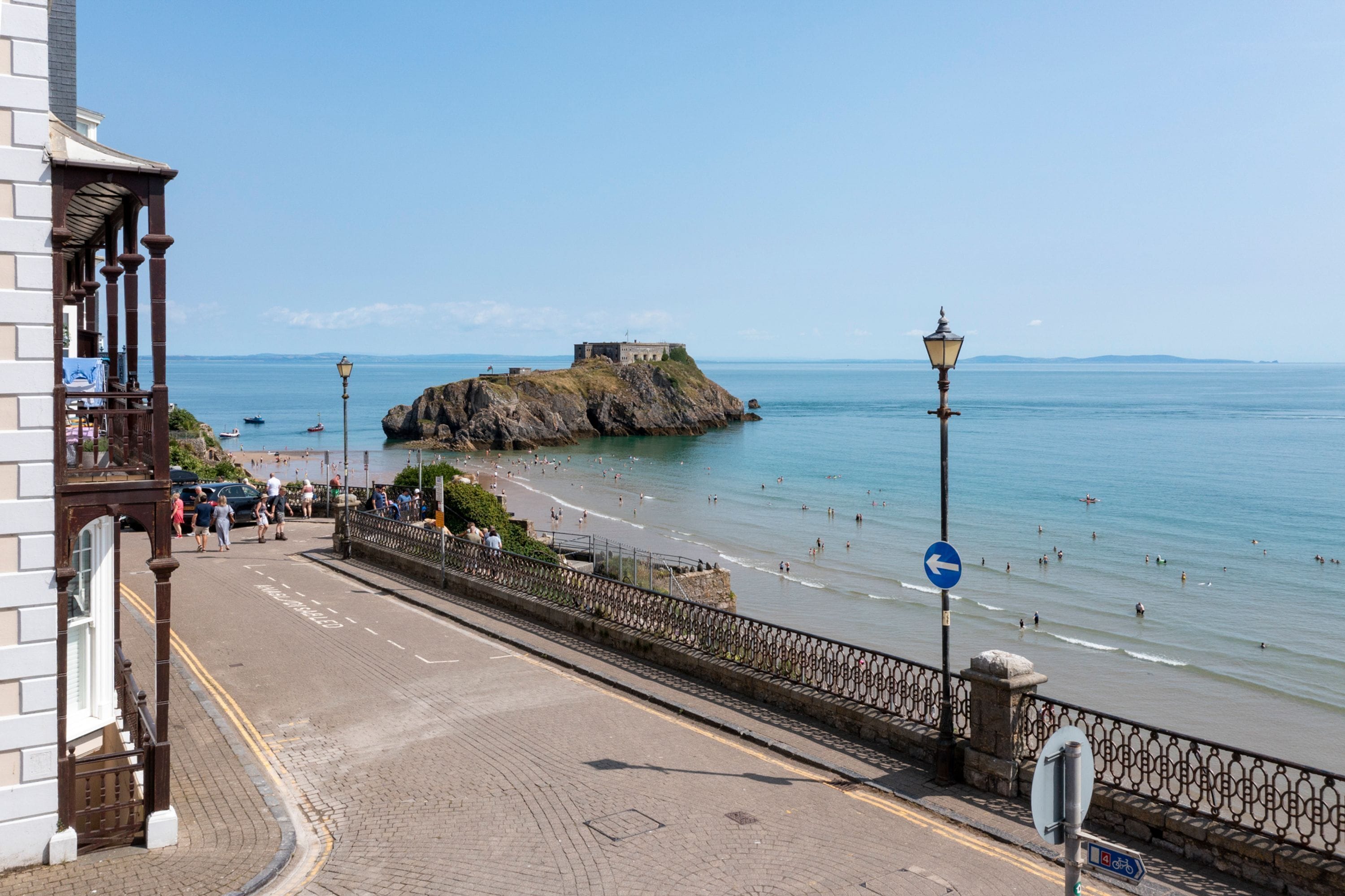 A coastal promenade with a stone balustrade, a beach, and a rocky outcrop with a building on top.