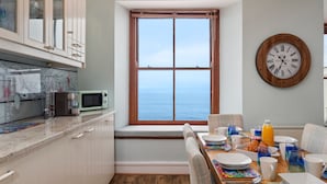 A kitchen with a view of the sea, a dining table set with breakfast, and a wall clock. - 6 Paragon Court - Sea Front Apartment (Tenby)