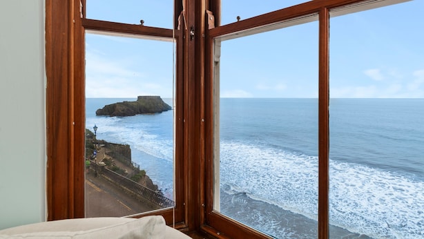 A room with a large window overlooking a coastal view, featuring a rocky outcrop and the sea. - 6 Paragon Court - Sea Front Apartment (Tenby)