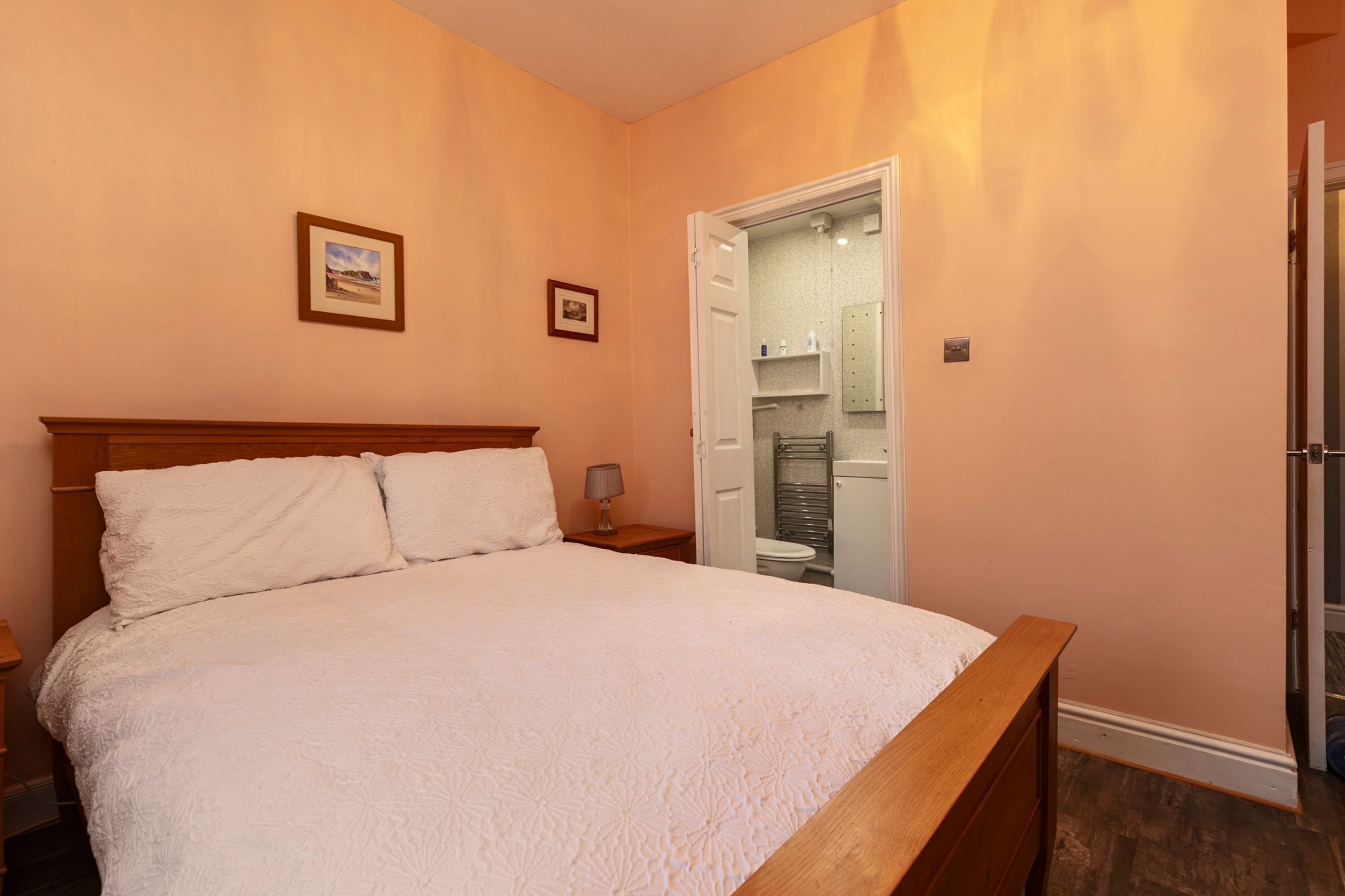 A bedroom with a large bed, wooden headboard, two framed pictures on the wall, and a bathroom visible through an open door.