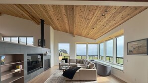 A modern living room with a fireplace, a sofa, and large windows offering a view. - Sea Legacy | New Construction, Ocean View, Hot Tub (Sea Ranch)