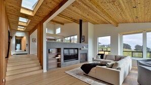 A spacious living area with a fireplace, wooden flooring, and large windows offering views of the outdoors. - Sea Legacy | New Construction, Ocean View, Hot Tub (Sea Ranch)
