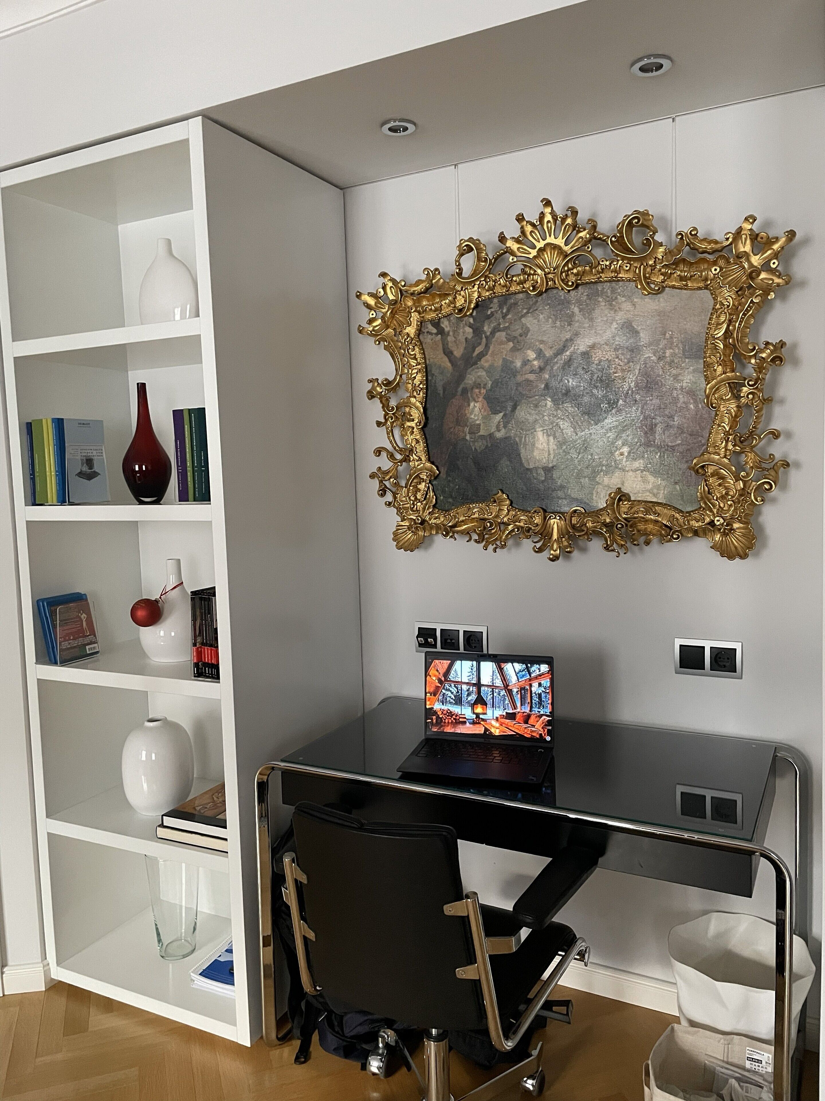 A modern office space with a glass desk, a laptop, a bookshelf with decorative items, and a framed painting on the wall.