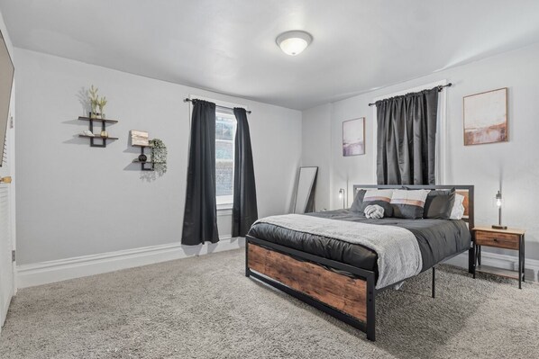 Quarto com cama, mesa de cabeceira, prateleira na parede com plantas e janela com cortinas.