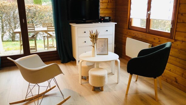 Een knus interieur met houten details, een witte tafel met een klein voetenbankje, een zwarte stoel, een witte schommelstoel en een televisie op een houten kast.