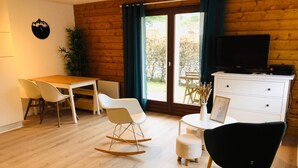 Een moderne woonkamer met een houten vloer, een eettafel met stoelen, een schommelstoel, een zwarte stoel, een witte kast en een televisie.