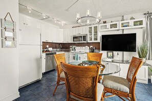 A modern kitchen with white cabinets, a dining table with wicker chairs, a large flat-screen TV, and a hanging light fixture. - Sunset Gulf‑Front Condo  Modern Coastal Interior, Steps to Sand (Fort Myers Beach)