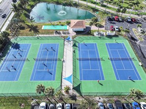 Two tennis courts with a small pond and a building in the background. - Sunset Gulf‑Front Condo  Modern Coastal Interior, Steps to Sand (Fort Myers Beach)