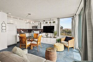 A modern kitchen with white cabinets, a dining area with wicker chairs, a flat-screen TV, and a large window with curtains. - Sunset Gulf‑Front Condo  Modern Coastal Interior, Steps to Sand (Fort Myers Beach)