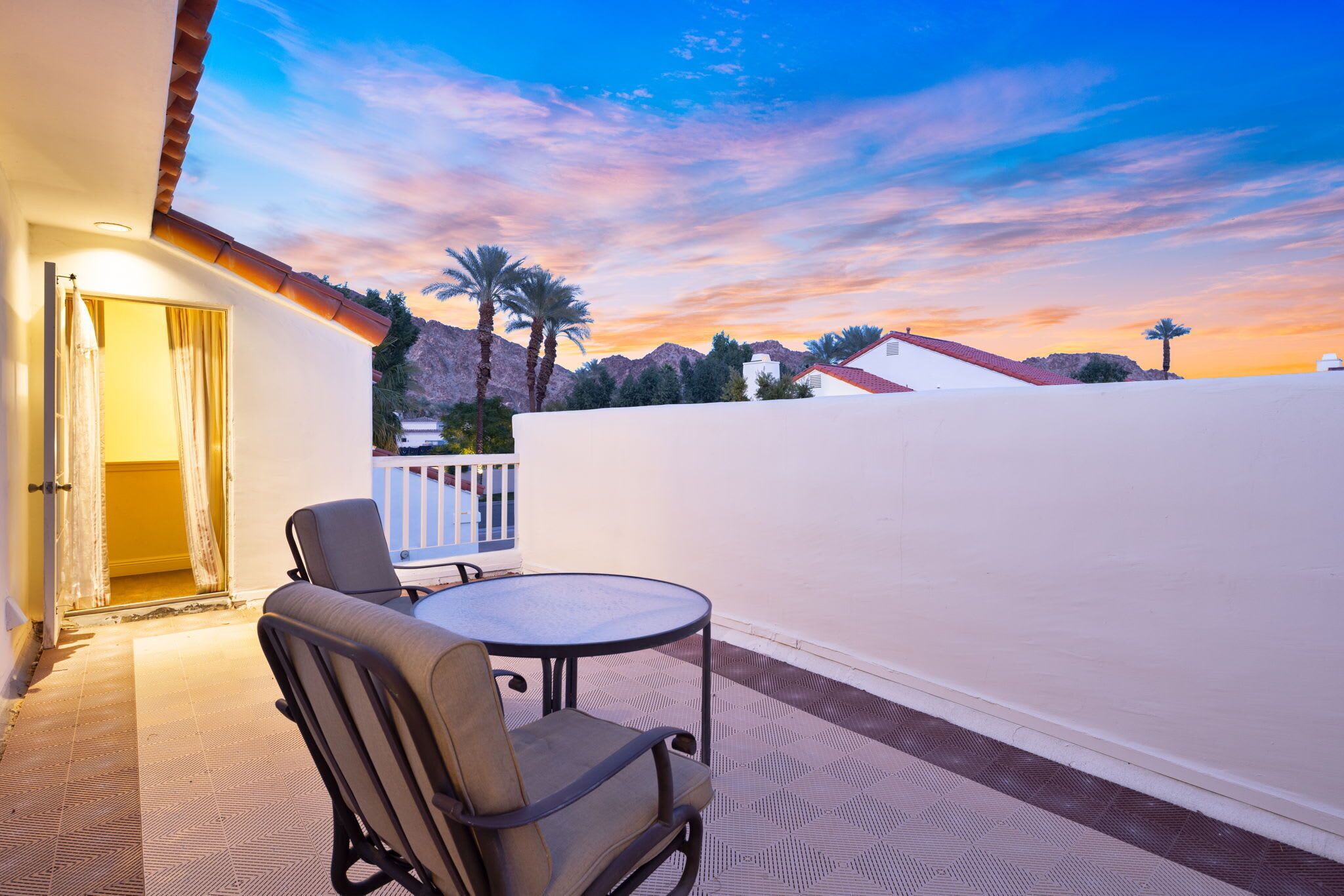 A balcony with a table and chairs, overlooking a mountainous landscape during sunset.