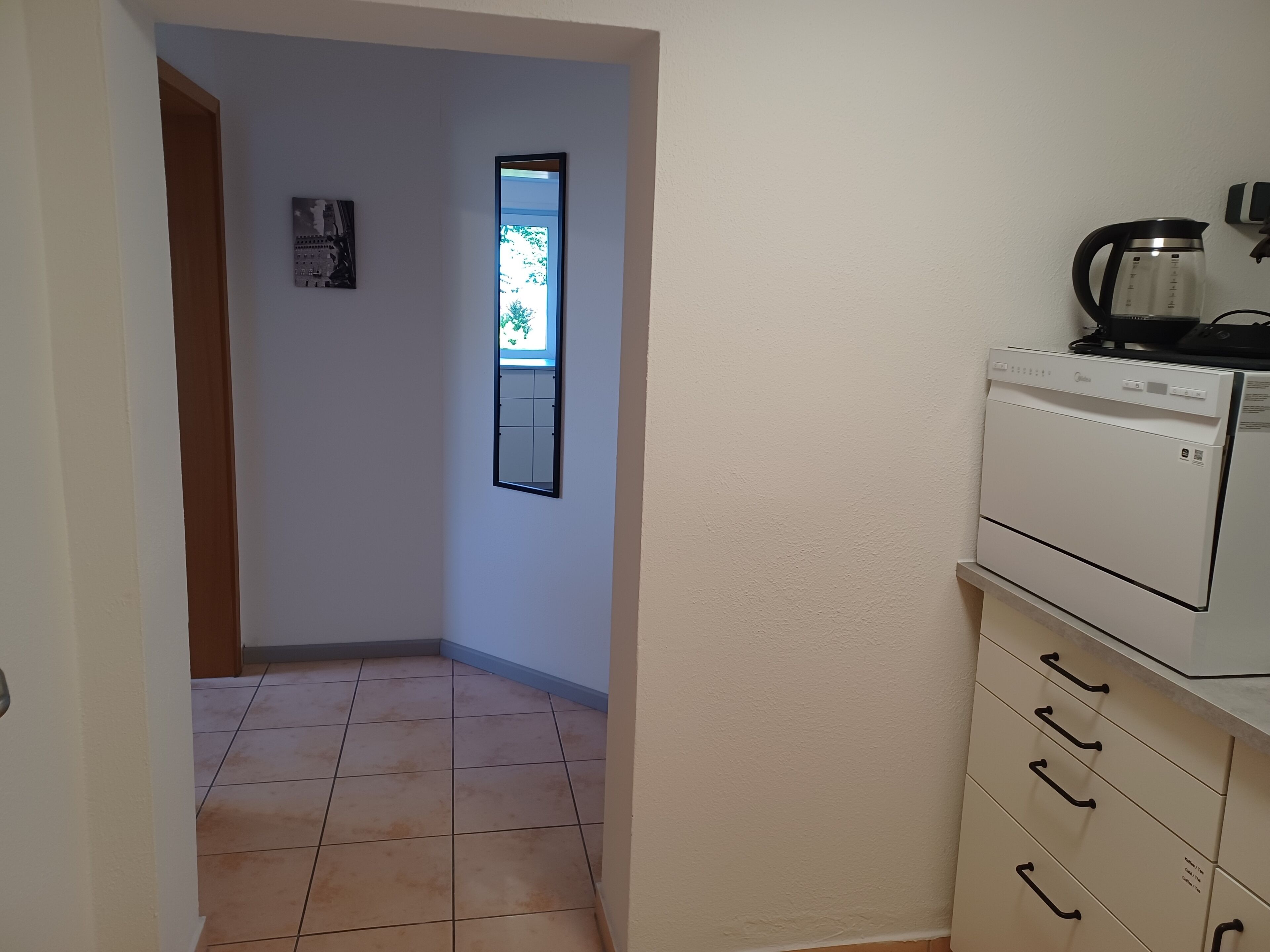 Un pasillo estrecho con suelo de baldosas, una ventana con vistas y una pequeña cocina equipada con cafetera y lavavajillas.