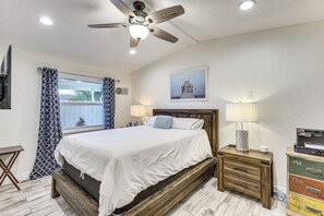 Ein Schlafzimmer mit Bett, Deckenventilator, Fenster mit Vorhängen, Nachttisch mit Lampe und einem Wandbild.