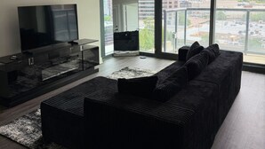 Una sala de estar moderna con un televisor grande, un sofá negro y vistas a un paisaje urbano a través de amplios ventanales.