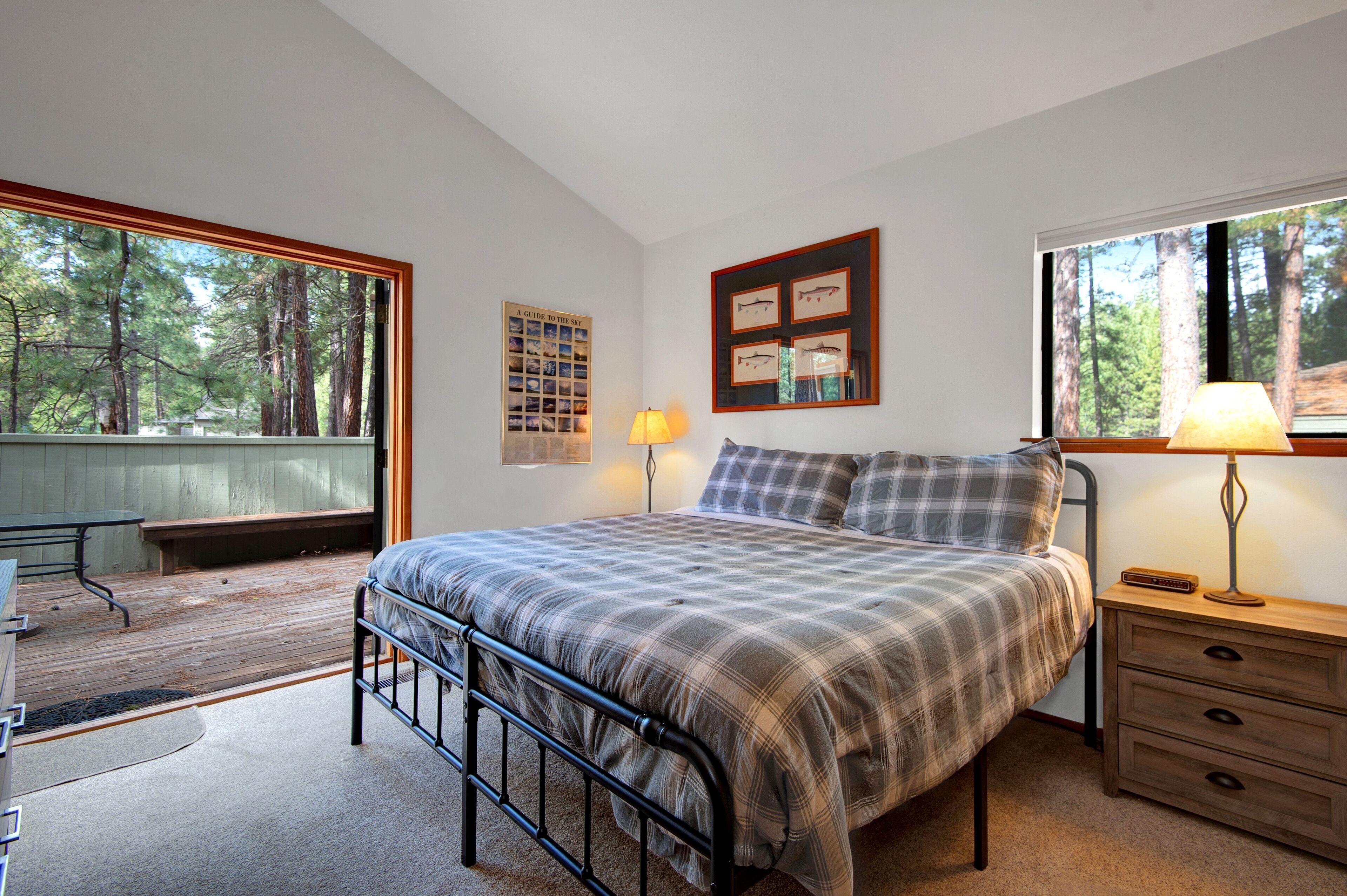 A bedroom with a bed, a nightstand, a lamp, a desk, and a window with a view of trees.