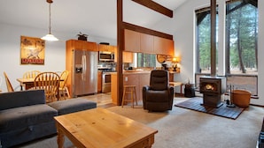 Ein gemütliches Wohnzimmer mit einer Couch, einem Holztisch und einem Kamin. Ebenfalls sichtbar sind eine Küche mit Holzschränken sowie ein Essbereich mit Tisch und Stühlen.