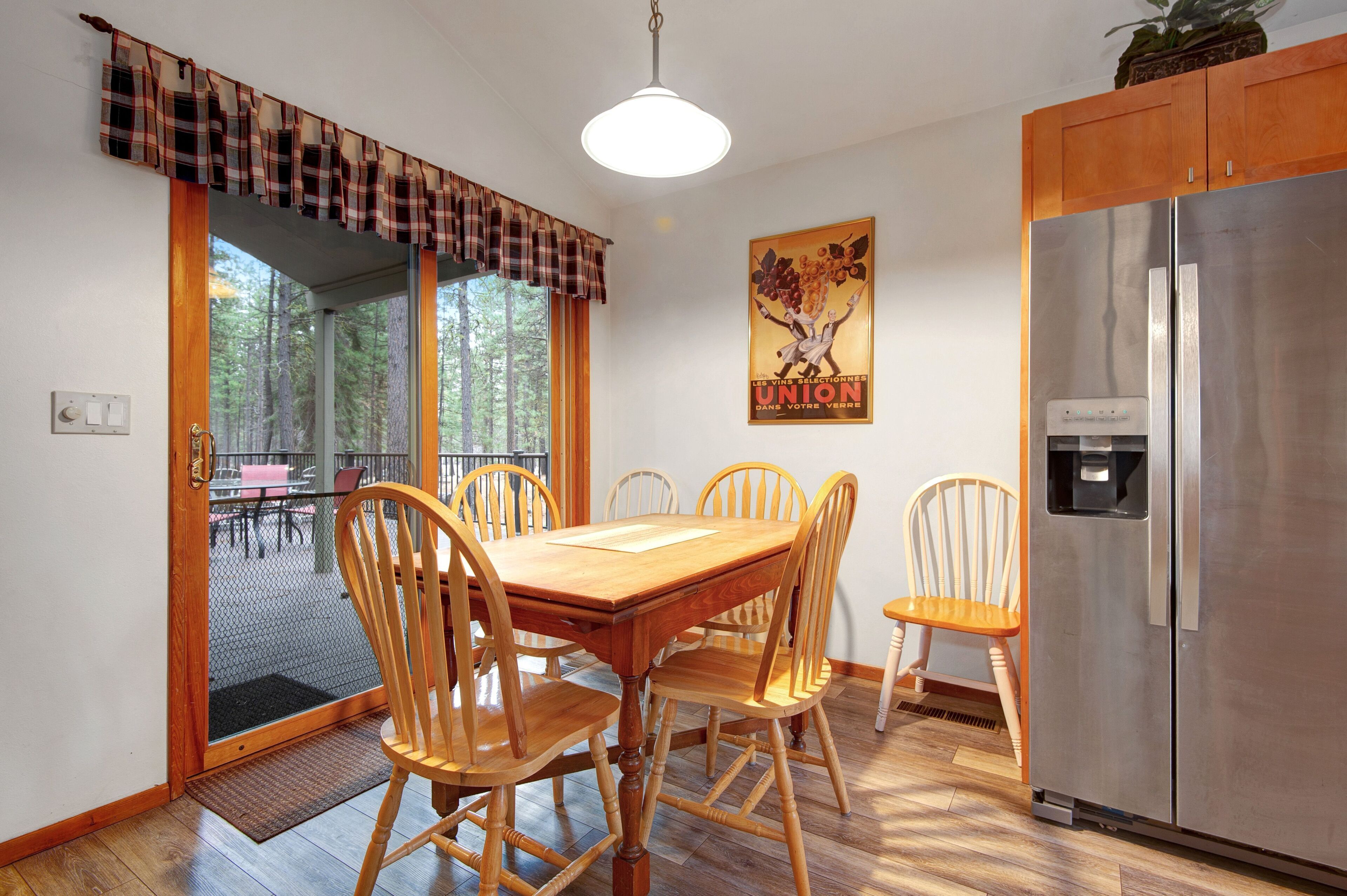 A dining area with a wooden table and chairs, a stainless steel refrigerator, and a window with a view of trees.