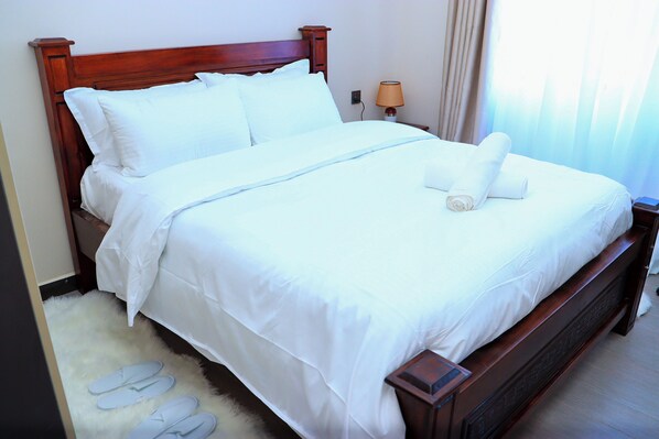A neatly made bed with white linens and a rolled towel, a wooden headboard, and a nightstand with a lamp. - Top Notch One-Bedroom Apartment (Nairobi)