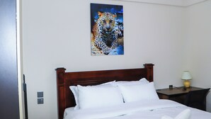 Ein Hotelzimmer mit einem hölzernen Bett, weißer Bettwäsche, einem Leopardenbild an der Wand, einem Nachttisch mit Lampe und einer Steckdose.