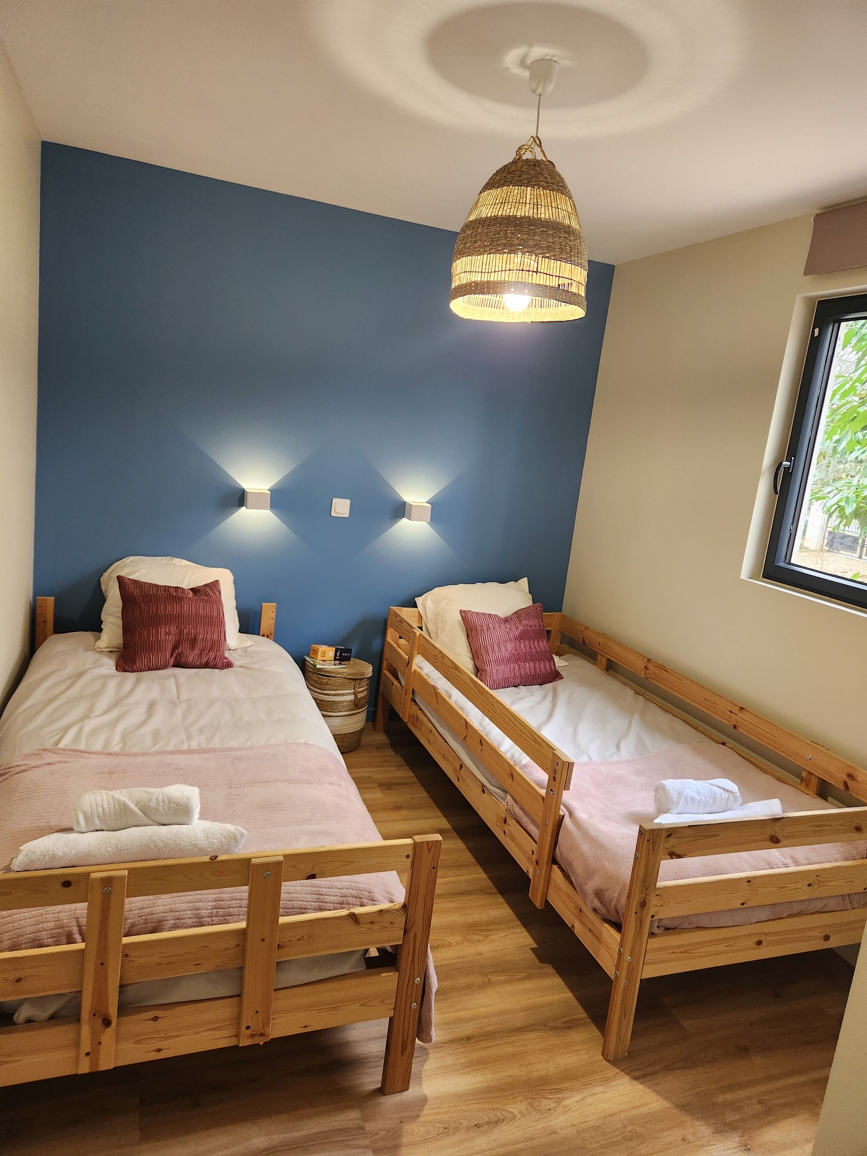 A room with two wooden bunk beds, a woven ceiling light, and a window with a view of greenery.