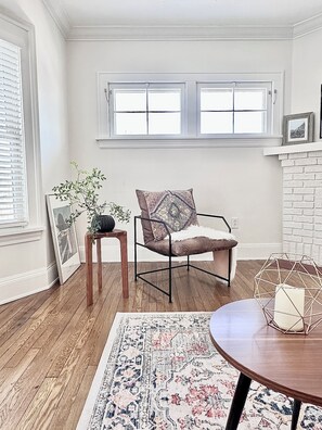 A room with a wooden floor, a patterned rug, a chair with a cushion, a small table with a geometric vase, and a framed picture on the wall.