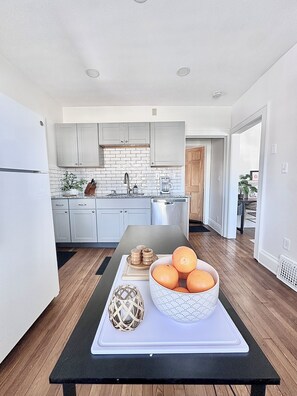A modern kitchen with white cabinets, a black countertop, and a wooden dining table with a bowl of oranges and decorative items.