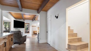 A spacious living area with a wooden ceiling, a flat-screen TV mounted on the wall, a grey sofa, and a wooden staircase. - The Herold's Shaver Lake Retreat | BBQ, Fire pit+ (Shaver Lake)