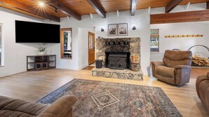 A spacious living room with a stone fireplace, wooden beams on the ceiling, a flat-screen TV, and a cozy seating area with a patterned rug. - The Herold's Shaver Lake Retreat | BBQ, Fire pit+ (Shaver Lake)