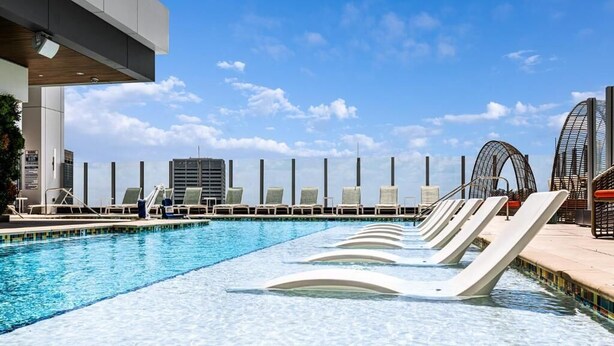 Una piscina con lettini e uno scivolo, circondata da edifici moderni e un cielo sereno.