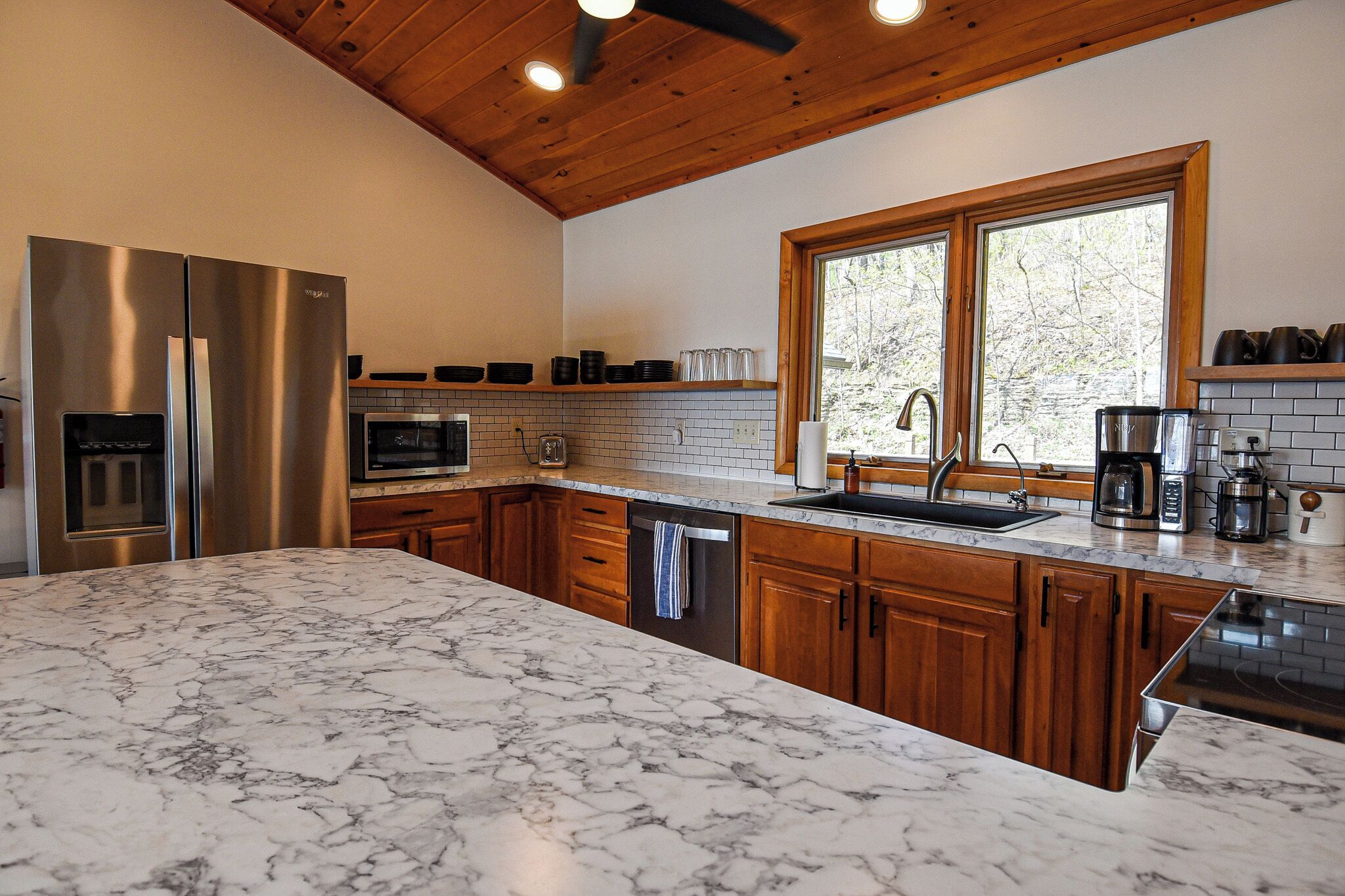 A kitchen with a marble countertop, stainless steel appliances, wooden cabinets, and a window with a view of trees.