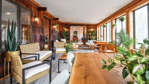Quarto espaçoso com paredes de madeira, grandes janelas e diferentes opções de assento, incluindo uma mesa de madeira e cadeiras de vime.