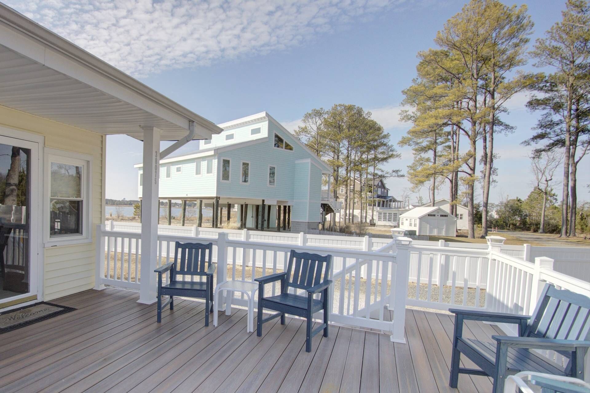 A wooden deck with a white railing, three dark blue Adirondack chairs, and a light blue house with a white porch.