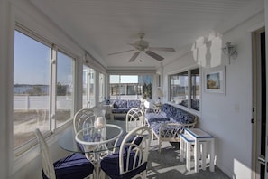A sunroom with a ceiling fan, a glass table, white metal chairs, patterned sofas, and large windows offering a view of the outdoors.