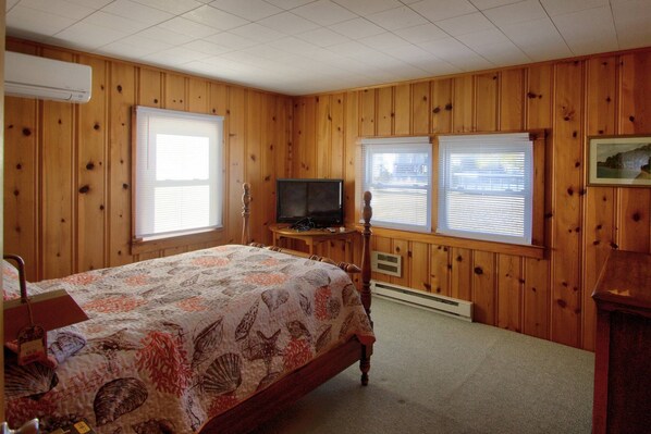 A bedroom with a wooden paneled wall, a bed with a floral bedspread, a television on a wooden stand, and two windows with blinds. - Pet Friendly Water Front Cottage Sleeping 8 with Kayaks and More Free Activities (Ocean View)