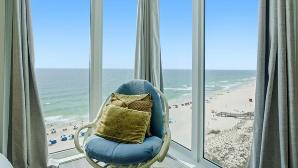 Een kamer met een groot raam met uitzicht op een strand, een rieten stoel met kussens, en een vergezicht op de oceaan en het strand.
