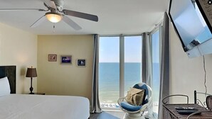 Hotelkamer met een bed, een plafondventilator, een televisie, een stoel met kussen, en uitzicht op de oceaan door grote ramen.