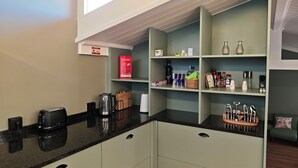 Eine moderne Küche mit integrierter Kaffeemaschine, Toaster und Wasserkocher auf der Arbeitsfläche. Regale mit diversen Küchenutensilien und eine kleine Sitzecke mit Stuhl und Kissen.