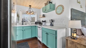Une cuisine avec des armoires de couleur turquoise, un fourneau blanc et un comptoir en bois. On y trouve une fenêtre avec des stores, un luminaire suspendu et une étagère fixée au mur, sur laquelle sont rangés des ustensiles de cuisine.