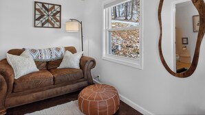 Une pièce avec un canapé en cuir marron, un pouf rond, un miroir fixé au mur, une lampe sur pied et une fenêtre donnant sur un paysage enneigé.