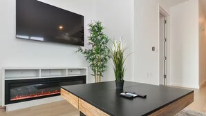 A modern living room with a flat-screen TV, a fireplace, a wooden coffee table, and two potted plants. - Suites at Dog Lane (Mansfield)
