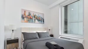 A modern bedroom with a large bed, a cityscape painting, and a large window. - Suites at Dog Lane (Mansfield)