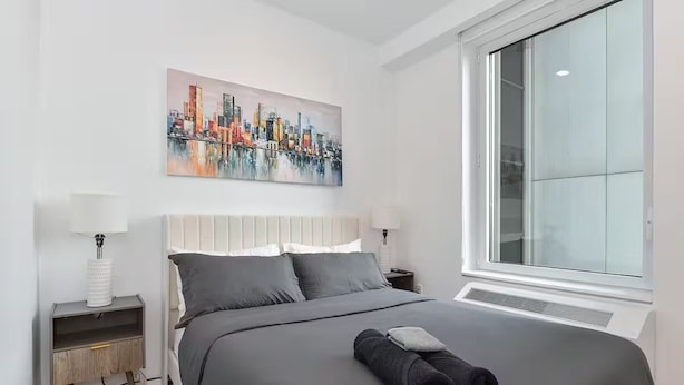 A modern bedroom with a large bed, a cityscape painting, and a large window. - Suites at Dog Lane (Mansfield)