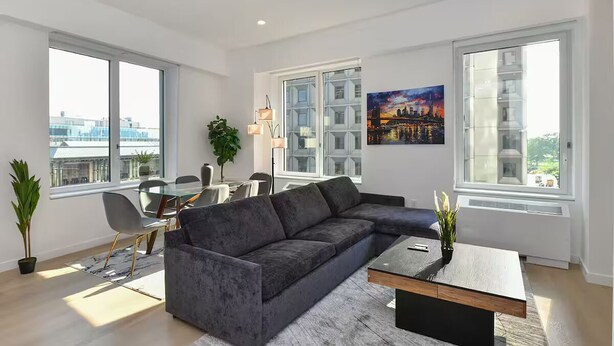 A modern living room with a sectional sofa, dining table, chairs, large windows, and a cityscape painting. - Suites at Dog Lane (Mansfield)